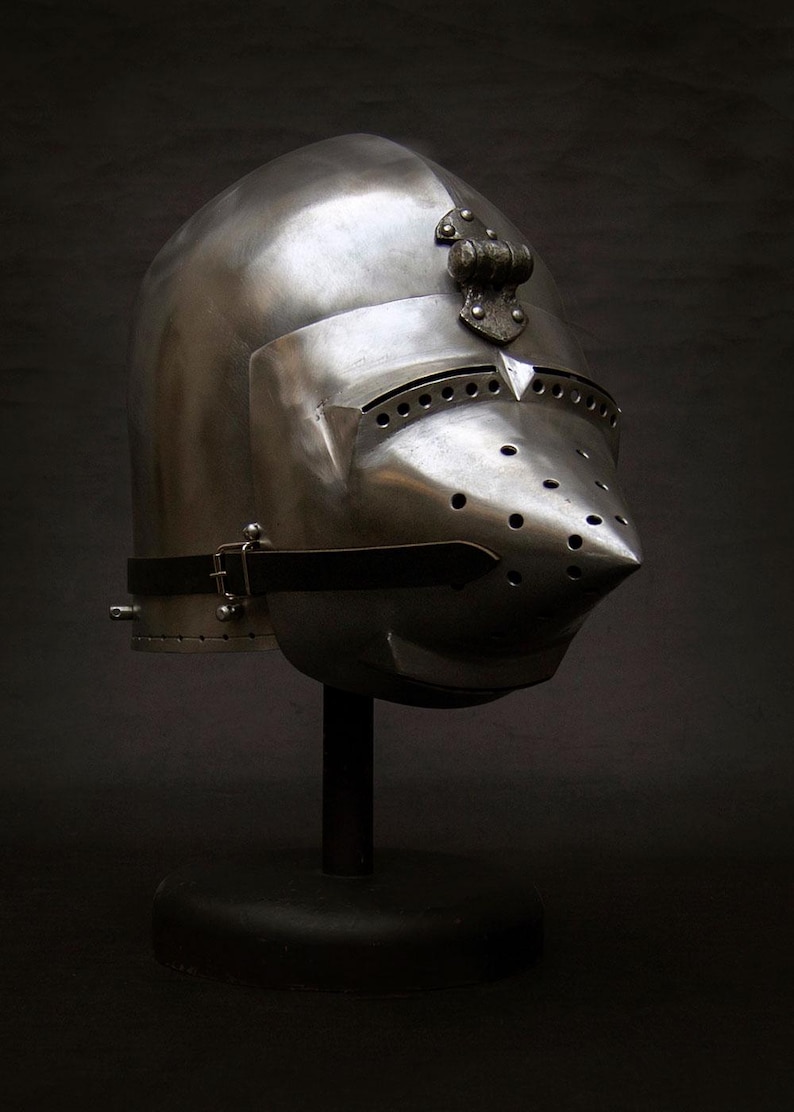 May include: A polished steel medieval helmet with a pointed visor and numerous ventilation holes. The helmet features a decorative element at the crown and a leather strap with a buckle. It is displayed on a black stand against a dark background.