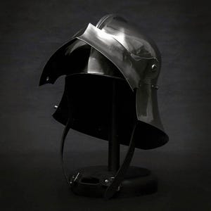 K&ouml;nnte beinhalten: Ein polierter Stahlhelm, wahrscheinlich f&uuml;r historische Nachstellungen oder Ausstellungen, vor dunklem Hintergrund. Der Helm hat eine abgerundete Krone, ein markantes Visier und einen Nackenschutz. Riemen sind sichtbar, was darauf hindeutet, dass er tragbar ist.