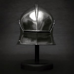 K&ouml;nnte beinhalten: Ein polierter, silberfarbener Helm, wahrscheinlich im mittelalterlichen oder Renaissance-Stil, auf einem schwarzen St&auml;nder ausgestellt. Der Helm hat einen markanten Mittelgrat und ein Visier. Der Hintergrund ist dunkelgrau und erzeugt einen dramatischen Kontrast.