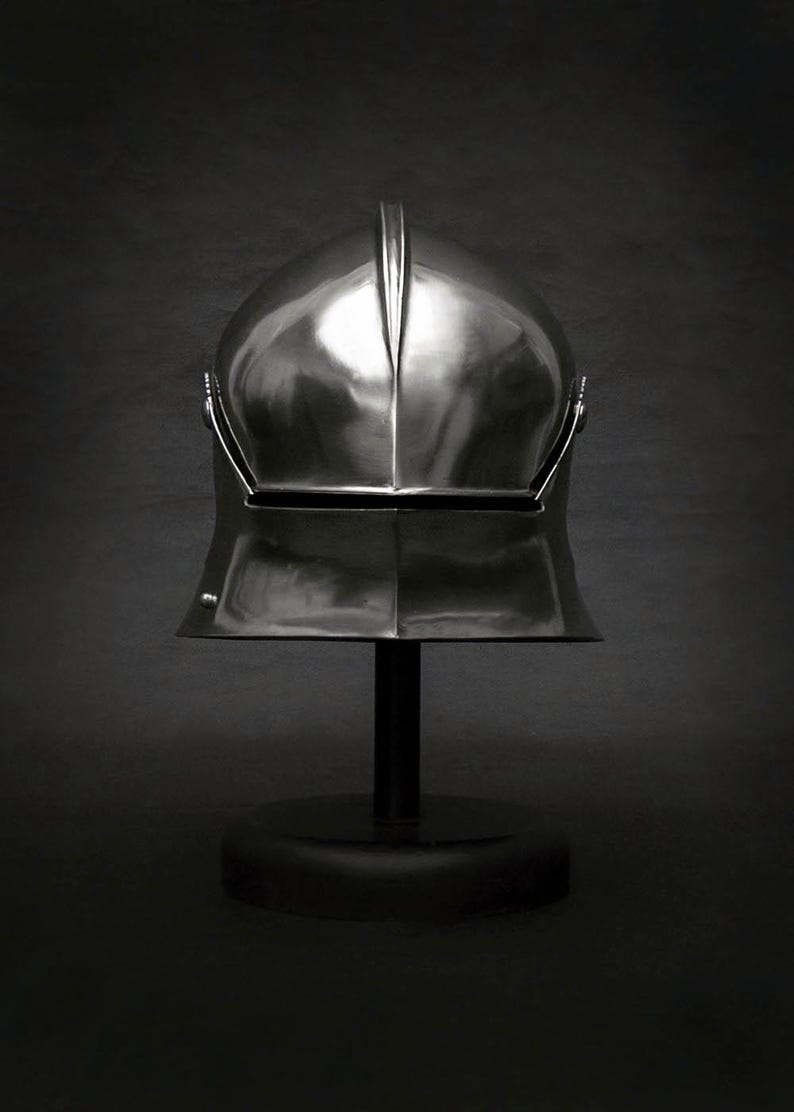 K&ouml;nnte beinhalten: Ein polierter, silberfarbener Helm, wahrscheinlich im mittelalterlichen oder Renaissance-Stil, auf einem schwarzen St&auml;nder ausgestellt. Der Helm hat einen markanten Mittelgrat und ein Visier. Der Hintergrund ist dunkelgrau und erzeugt einen dramatischen Kontrast.