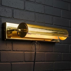 Op de afbeelding: Een messingkleurige wandlamp met een cilindrische kap en een rechthoekige basis, gemonteerd op een donkere bakstenen muur. Het licht straalt warmte uit. Een stijlvolle toevoeging aan elk interieur.