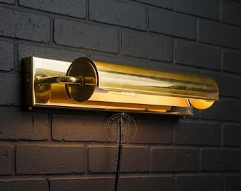 May include: A brass-colored wall-mounted light fixture with a cylindrical shade and a rectangular base. The light is mounted on a dark brick wall. The fixture has a warm, inviting glow.