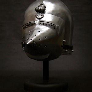 May include: A polished steel medieval helmet with a rounded top and a pointed visor. The helmet has numerous small holes for ventilation and is mounted on a black stand. The helmet is a historical artifact.
