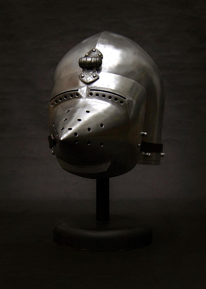 May include: A polished steel medieval helmet with a rounded top and a pointed visor. The helmet has numerous small holes for ventilation and is mounted on a black stand. The helmet is a historical artifact.