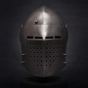 Op de afbeelding: Een middeleeuwse stalen helm met een puntige bovenkant en een frontplaat met talrijke kleine ventilatiegaten. De helm heeft een donkere, metalen afwerking en staat tegen een donkere achtergrond.