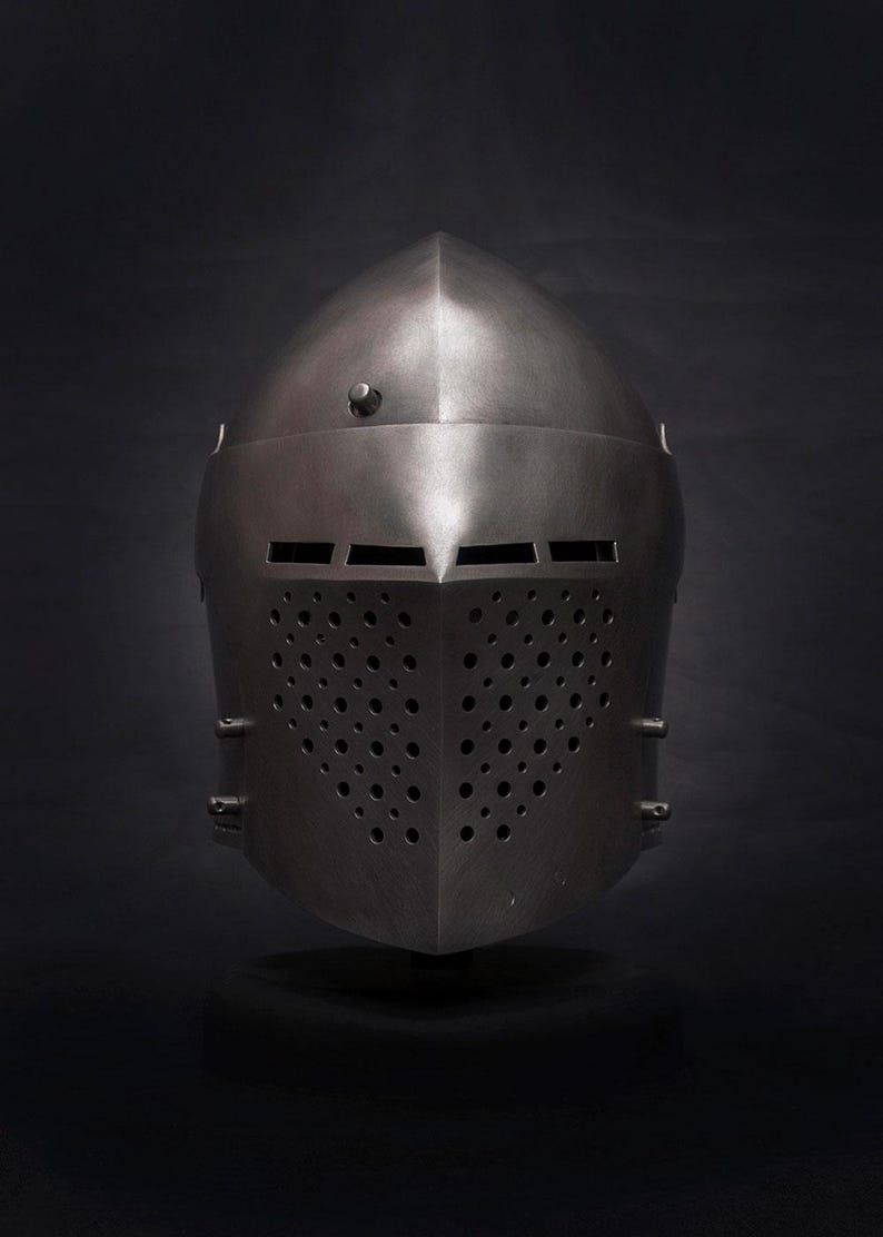 Op de afbeelding: Een middeleeuwse stalen helm met een puntige bovenkant en een frontplaat met talrijke kleine ventilatiegaten. De helm heeft een donkere, metalen afwerking en staat tegen een donkere achtergrond.