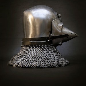 May include: A polished metal helmet with a pointed visor and a chainmail neck guard. The helmet has a dark leather strap and buckle. The medieval-style helmet is set against a dark background.