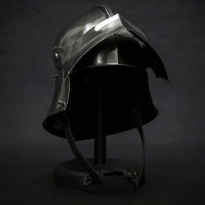 K&ouml;nnte beinhalten: Dunkler Metallhelm mit gebogenem Visier und genieteten Details, ausgestellt auf einem schwarzen St&auml;nder. Der Helm hat eine glatte, reflektierende Oberfl&auml;che und Lederriemen. Die Gesamt&auml;sthetik deutet auf historische R&uuml;stung hin.
