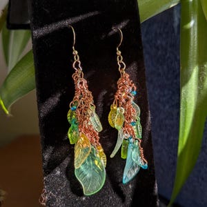 May include: A pair of handcrafted earrings featuring a cascade of green and yellow leaf-shaped beads, accented with small blue beads and copper wire. The earrings are displayed against a black backdrop, showcasing their intricate design and vibrant colors.