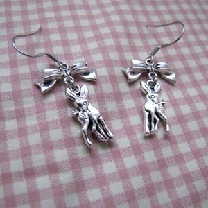 May include: A pair of silver earrings featuring a small deer charm and a bow. The earrings are on a pink and white gingham background.