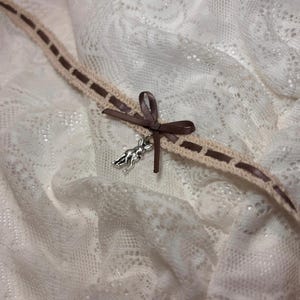 Puede incluir: Un collar gargantilla de crochet marrón y beige con un dije de plata de un ciervo. El collar está atado con un lazo de cinta de satén marrón.