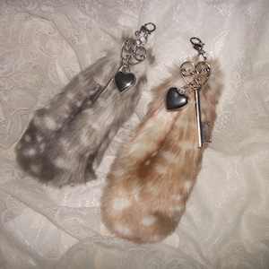 May include: Two fluffy keychains with silver keys and heart charms. The keychains are made of faux fur with a spotted pattern. One keychain is gray and white, the other is brown and white.