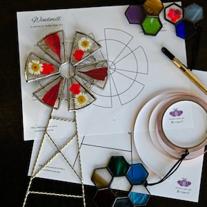 May include: A DIY stained glass windmill kit with a pattern, copper foil tape, and a variety of glass pieces. The kit includes a template for a windmill with eight sections, each with a different design. The glass pieces are in various colors, including red, green, blue, and purple. The kit also includes a pair of tweezers and a small pair of scissors.