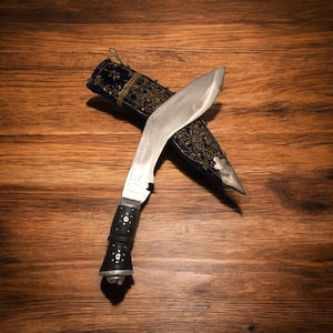 May include: A silver-colored knife with a black handle and a blue and gold embroidered sheath. The knife is curved and has a pointed tip. The sheath has a decorative design.