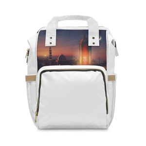 Puede incluir: Mochila blanca con una imagen colorida de una mezquita y una linterna al atardecer. La mochila tiene un bolsillo frontal grande, bolsillos laterales y un asa superior. Cremalleras y detalles dorados.
