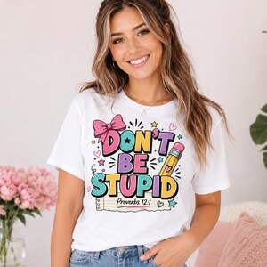 以下が含まれることがあります： 「DON'T BE STUPID」および「Proverbs 12:1」というテキストがプリントされた、カラフルなグラフィックデザインの白い半袖Tシャツ。デザインには、ピンクの水玉模様のリボン、黄色い鉛筆、パステルカラーの小さな星やハートの形が含まれています。