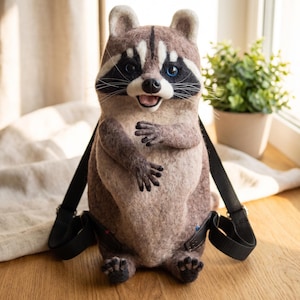 May include: A needle-felted raccoon backpack featuring a textured brown and grey body with a white chest and face mask. The raccoon has black eye patches, blue eyes, a black nose, and an open mouth. It includes black adjustable shoulder straps.
