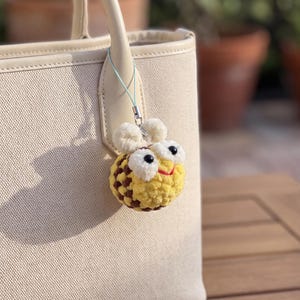 May include: A cream-colored canvas tote bag with a crochet bee charm attached to the handle. The bee is round with yellow and brown stripes, white fuzzy wings, large black eyes, and a small red stitched mouth, hanging from a light blue cord.