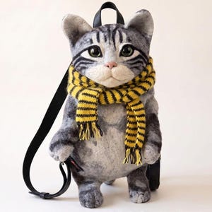 Puede incluir: Una mochila de gato hecha a mano en lana con forma de gato atigrado gris, ojos verdes grandes y una bufanda de punto a rayas amarillas y negras. La mochila tiene correas negras, mide 54 cm de alto y 34 cm de ancho. Texto: HANDMADE Wool CAT BACKPACK.