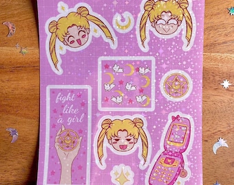 May include: A pink and white sticker sheet with a Sailor Moon theme. The sheet includes images of Sailor Moon, her signature crescent moon, a cell phone, a star, and the phrase "fight like a girl".