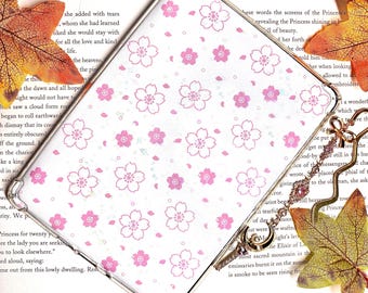 May include: A clear phone case with a pink and white floral pattern. The case is sitting on top of an open book with a brown cover. The book has the title "Tales of Japan" and the page number "74".