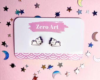 May include: A pair of white and black bunny stud earrings on a white card with the text "Zero Art". The card has a pink border with a wave pattern. The background is pink with star and moon-shaped confetti.