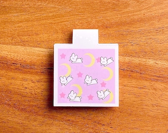 May include: A pink bookmark with a white border and a repeating pattern of white bunnies and yellow crescent moons on a pink background. The bookmark has a white tab at the top.