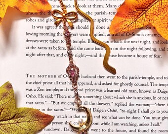 May include: A gold bookmark with a pink and white crystal charm and a bow. The bookmark is shaped like a person and is resting on a page of a book. The text on the page reads "Then the folk were afraid, and left the re r; and the mother of O-Sono's husband sa things; and O-Sono was much attached come back to look at them. Many de aniess the things robes and girdle temple, It was agreed thetras should the parish-tem er spirit w lowing morning the Awers were dresses were taken to temple. Bu done a bie. So o. optied; anu all of O-Sono's ornameı. came back the next night, and look on the night following, and t puse became a house of fear. at the tansu as before. And she came back night after that, and every night; and the in the tansu." "Well, THE MOTHER OF O-s husband then went to the parish-temple, and tol the chief priest all that hadaappened, and asked for ghostly counsel. The temp was a Zen temple; and the head-priest was a learned old man, known as Daige Oshō. He said: "There must be something about which she is anxious, in or nea that tansu."-"But we emped all the drawers," replied the woman; "there i d Daigen Osho, "to-night I shall go to your and see what can be done. You must give Joom while I am watching, unless I call." went to the house, and found the room there alone, reading the sûtras; and nothing the Rat. Then the figure of O-Sono suddenly su. Her face had a wistful look; and she kept ep watch in that rod person shall ent sundown, Daige nade ready for him. He rema appeared until after the Hour outlined itself in front of the her eyes fixed upon the tansu."
