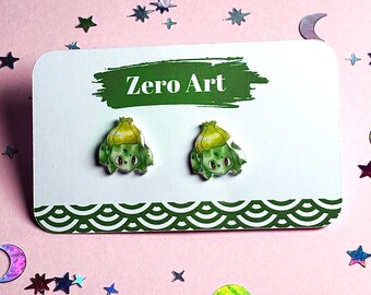May include: A pair of green and yellow Bulbasaur earrings on a white card with the text "Zero Art". The card has a green decorative border and is surrounded by star and moon-shaped confetti on a pink background.
