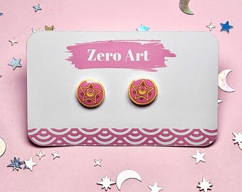 May include: Two round stud earrings with a pink and gold design featuring a star and crescent moon. The earrings are displayed on a white card with the text "Zero Art" and a pink patterned border, set against a pink background with star and moon decorations.