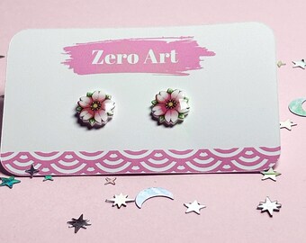 May include: A pair of floral stud earrings on a white card with the text "Zero Art" in pink. The earrings feature pink and white flower designs with green leaves. The card has a pink patterned border and is on a pink surface with star and moon-shaped confetti.