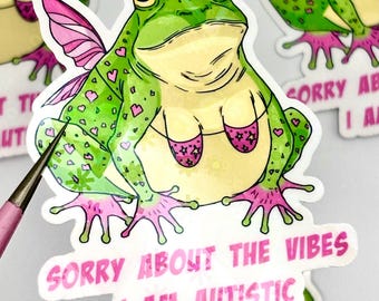 May include: A sticker featuring a whimsical green frog with pink fairy wings and a bikini. The frog has heart-shaped spots and the text "SORRY ABOUT THE VIBES I AM AUTISTIC" in pink.