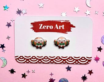 May include: A pair of owl-shaped stud earrings on a white card with the text "Zero Art". The earrings are brown, orange, and green, with a cartoon-like design. The card has a red border and is on a pink background with star and moon-shaped decorations.