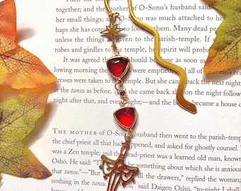 May include: A gold bookmark with red glass accents shaped like hearts. The bookmark is shaped like a sword and has a star design at the top. The bookmark is resting on a page of a book with text about a temple and a priest.