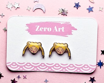 May include: A pair of stud earrings featuring a cartoon character with blonde hair and a smiling face. The earrings are displayed on a white card with the text "Zero Art" and a pink border, set against a pink background with star and moon-shaped decorations.