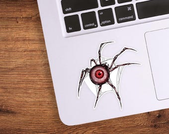 May include: A sticker featuring a stylized spider with an eyeball in the center. The spider is black and red, with a red eyeball. The sticker is on a white surface, with a wooden table in the background.