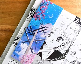 May include: A blue and pink bookmark with a glittery finish. The bookmark features a cityscape with a power line and cherry blossoms. The bookmark also has a crescent moon and stars.