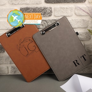 May include: Two personalized leather clipboards. One is brown with a floral design and the initials "JG" with the name "Joshua Gonzalez" underneath. The other is gray with the initials "RT" in a vertical design.