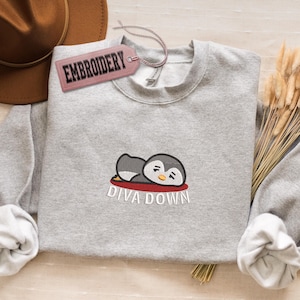 May include: A light gray sweatshirt with a crew neck features an embroidered design of a cartoon penguin resting on a red surface. The text "DIVA DOWN" is embroidered below the penguin. A pink tag that says "EMBROIDERY" is attached to the sweatshirt.
