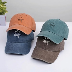 May include: Four baseball caps in shades of orange, blue, teal, and brown are stacked. Each cap features a sun and wave design embroidered in white. The caps have a classic, unstructured fit.