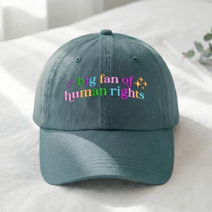 May include: A teal baseball cap with the embroidered text "big fan of human rights" in colorful lettering. The hat has a curved brim and a button on top. The text includes a star graphic.