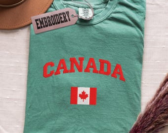 Puede incluir: Una camiseta verde azulado con la palabra "CANADA" en rojo, con letras arqueadas y una bandera canadiense. La camiseta tiene una etiqueta que dice "EMBROIDERY". Un sombrero marrón es visible en la esquina superior izquierda.