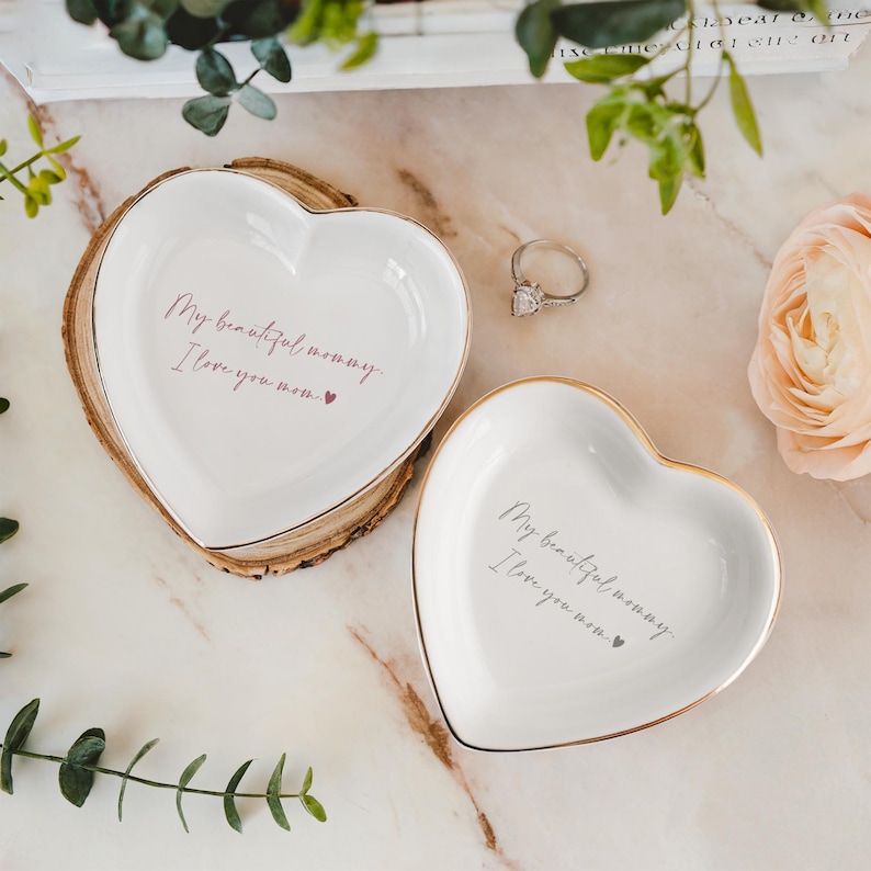 May include: Two white heart-shaped trinket dishes with gold rims. One dish rests on a wooden slice. The text "My beautiful mommy, I love you mom" is printed on each dish. A silver ring with a large stone is also visible.