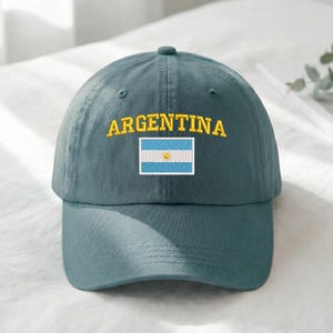 May include: A teal baseball cap with the word "ARGENTINA" embroidered in yellow above a small Argentinian flag. The flag is a light blue and white horizontal stripe design with a yellow sun in the center.