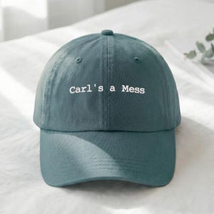 May include: A teal baseball cap with the text "Carl's a Mess" embroidered in white. The cap has a curved brim and a button on top. The hat is made of a soft fabric.