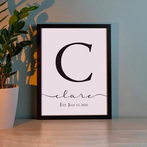 May include: A framed print featuring a large black letter "C" on a light pink background. Below the letter, the name "clare" is written in cursive, with the text "Est. July 13, 2025" underneath. The frame is black.