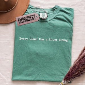 Può includere: Una maglietta verde acqua con la frase "Every Cloud Has a Silver Lining" stampata in bianco. Sono visibili un cappello marrone e un'etichetta con la scritta "EMBROIDERY". Un mazzo di fiori secchi si trova nell'angolo in basso a destra.