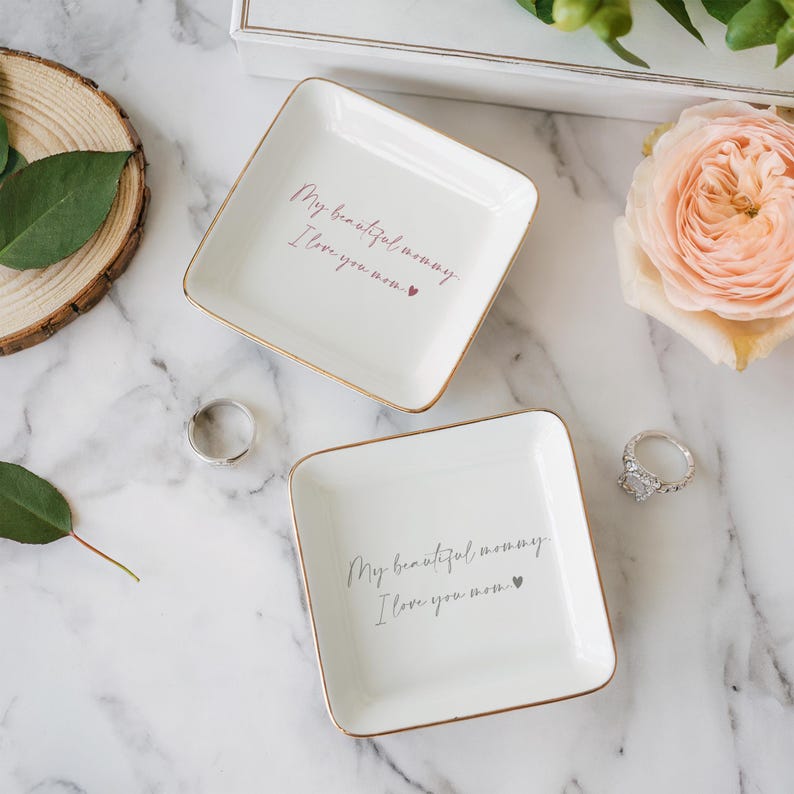 May include: Two white square trinket dishes with gold trim, each inscribed with "My beautiful mommy, I love you mom." A silver ring and a diamond ring are next to the dishes. A pink rose and greenery are in the background.