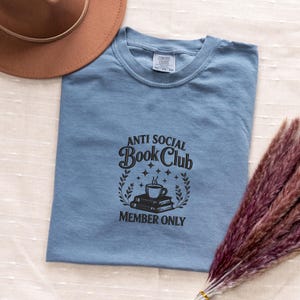 May include: A light blue t-shirt with the text "ANTI SOCIAL Book Club MEMBER ONLY" in black. The design includes a stack of books, a coffee cup, and decorative elements. A brown hat and dried flowers are in the background.