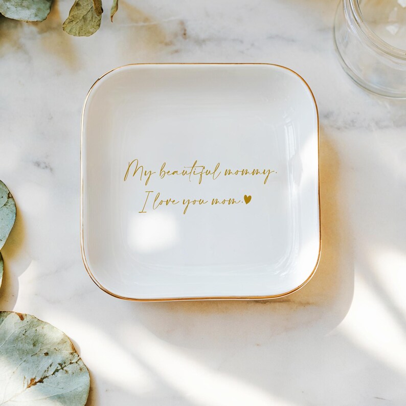 May include: White square trinket dish with gold trim. The dish has the text "My beautiful mommy. I love you mom." in gold script. A small gold heart is at the end of the text. The dish is on a white marble surface.